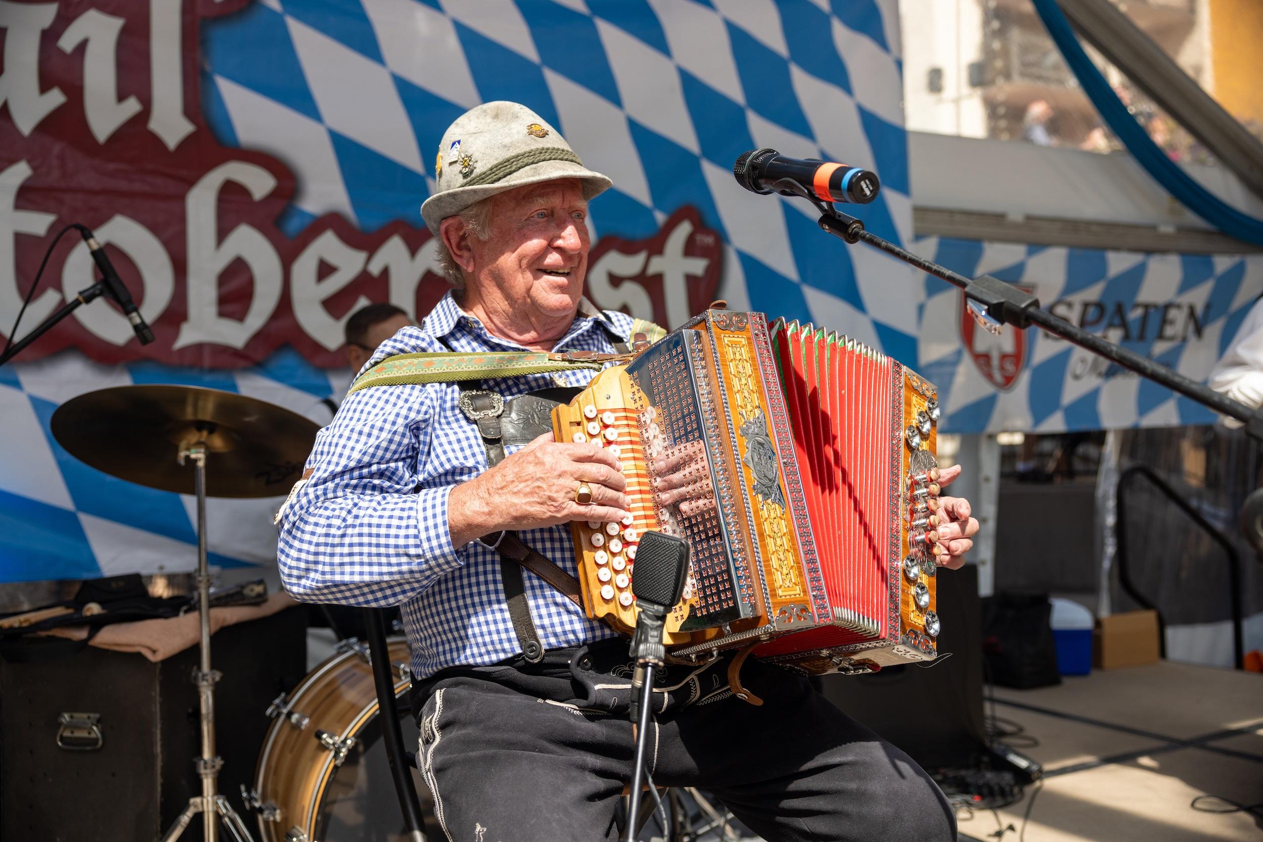 Photo shows a man seated on stage playing an accordion. He is in front of a blue and white checkered background and he is wearing a blue and white checkered shirt, lederhosen and a Bavarian hat. He is smiling.