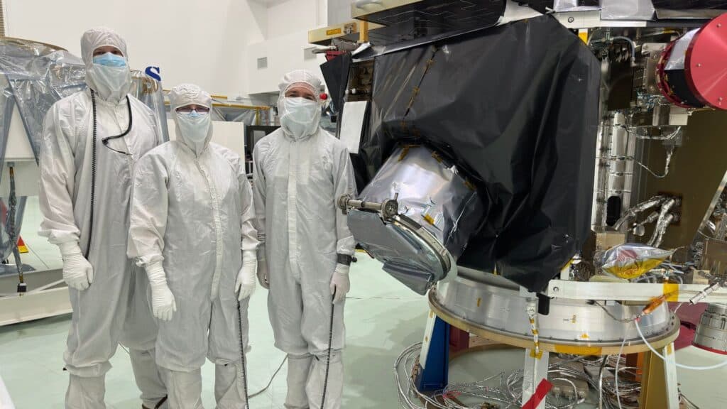Three people wearing white suits are standing in front of a black object.