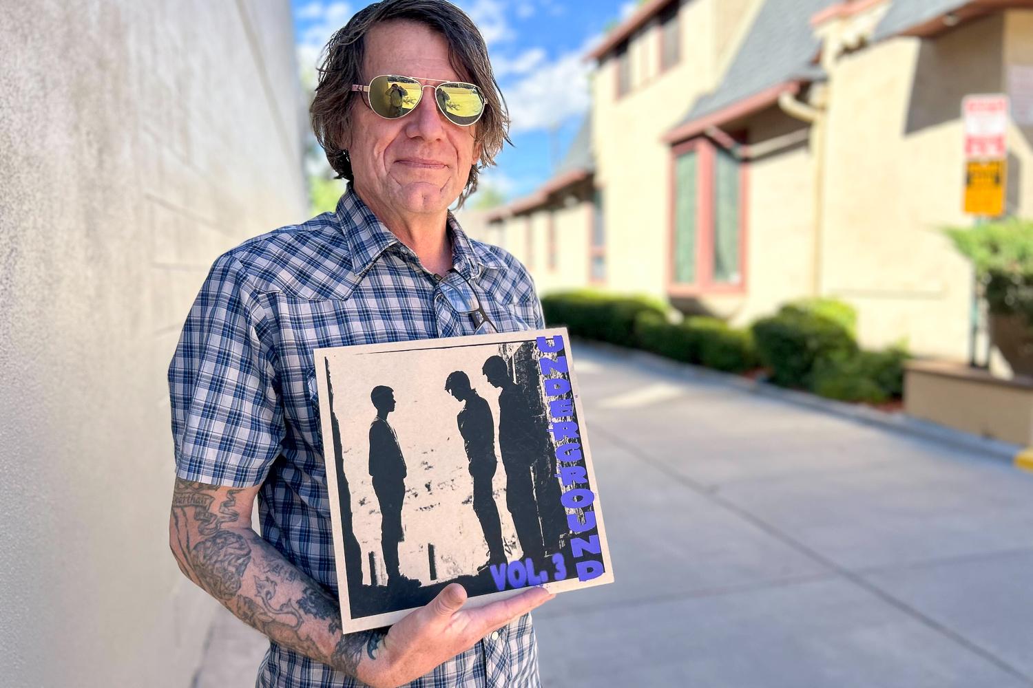 Danny Stewart, a man, stands outside the KRCC building in downtown Colorado Springs. He holds a record called Colorado Springs Underground Volume 3. He wears yellow mirrored sunglasses and a plaid shirt.
