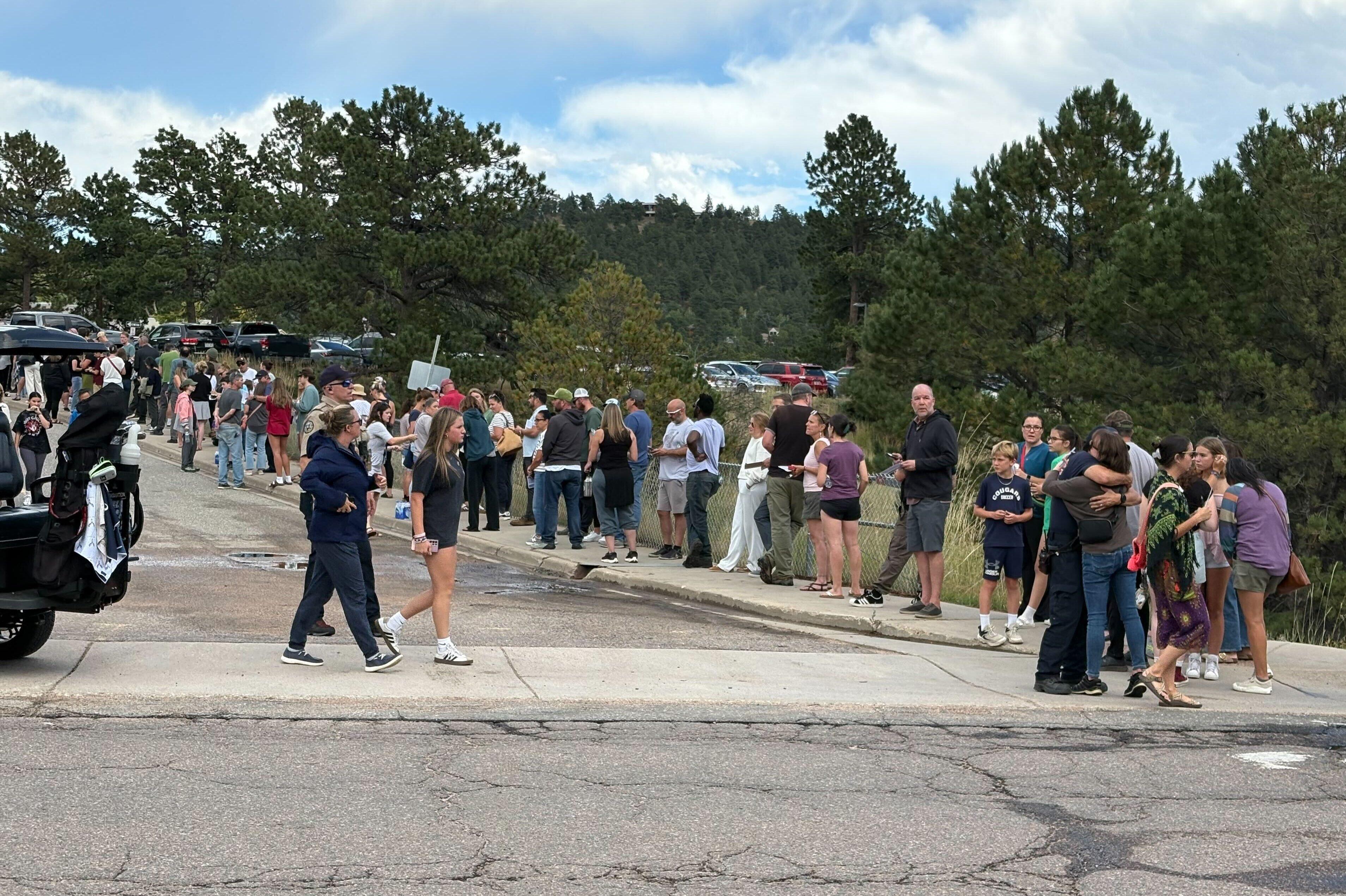 A large line of people stand and walk along a sidewalk near parked cars and trees, some talking, hugging, or waiting, on a partly cloudy day.