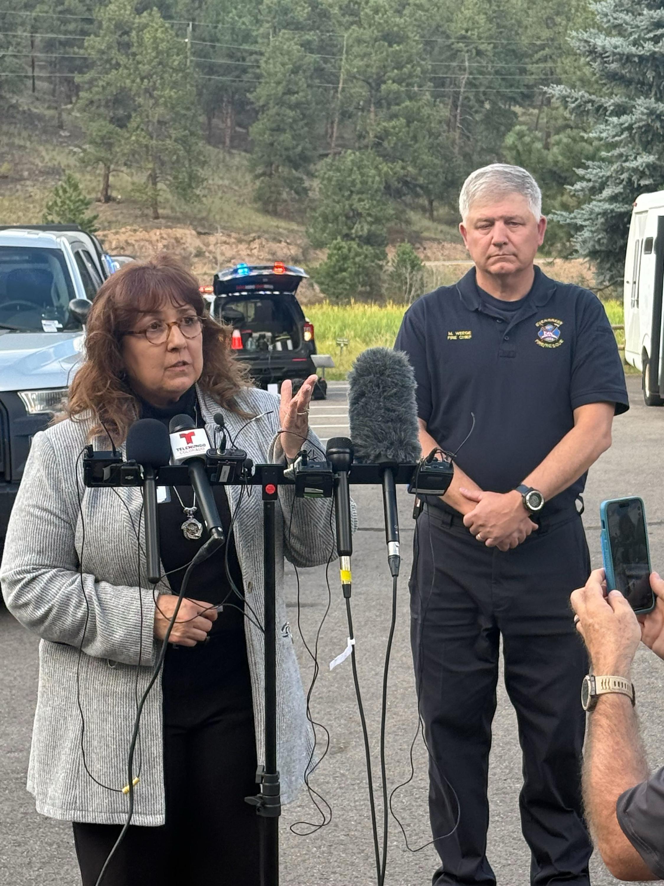 A woman speaks in front of a microphone array. She has glasses on and wears a gray cardigan over a black shirt and pants. Standing behind her is a man with grey hair in a firefighting polo. The two are addressing the press in a parking lot with trees in the background.