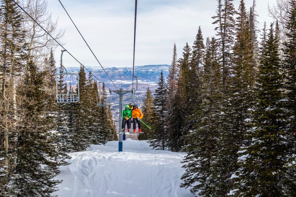 Two skiers ride an old, two-person chairlift through a stand of pine trees at Powderhorn Mountain Resort.