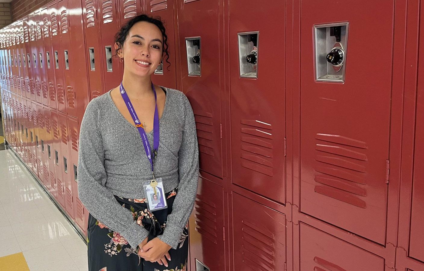 A woman in a gray v-neck top and floral print skirt poses smiling in front of rows of red school lockers.