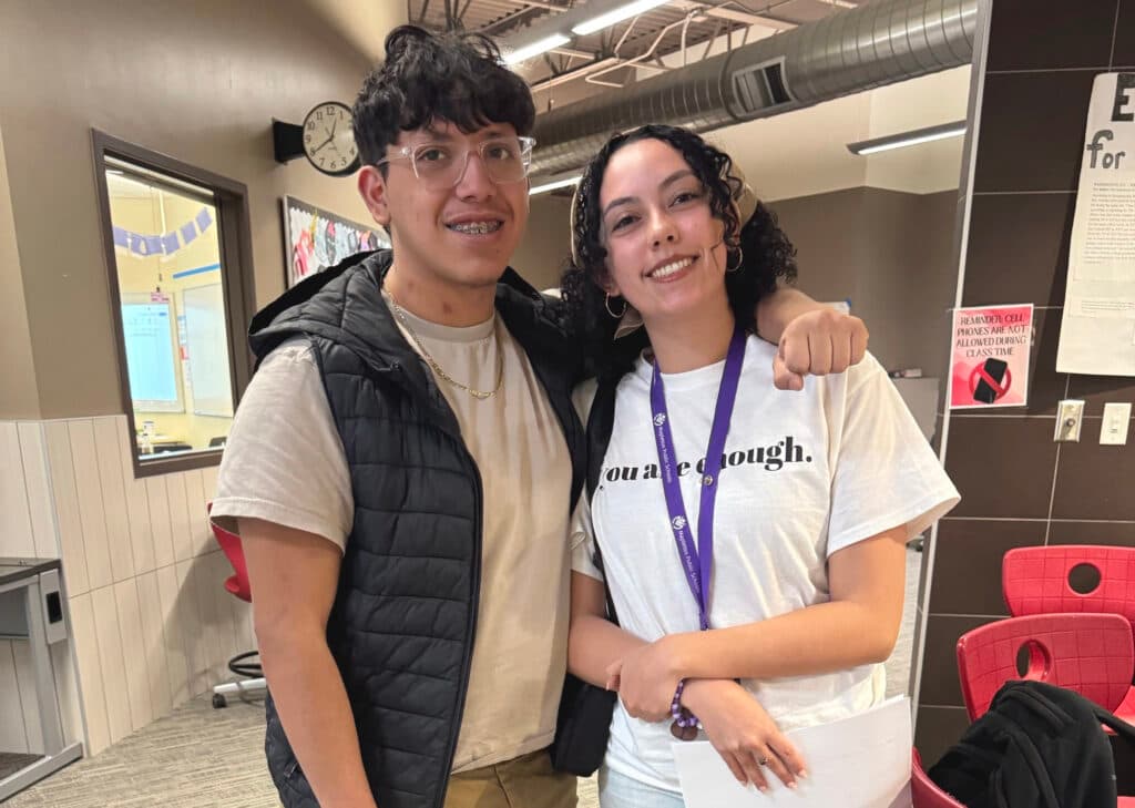 A young boy in clear-framed glasses and braces puts his arm around a woman to his left. She wears a lanyard and a T-shirt that says &quot;You are enough.&quot; Both are looking at the camera and smiling.