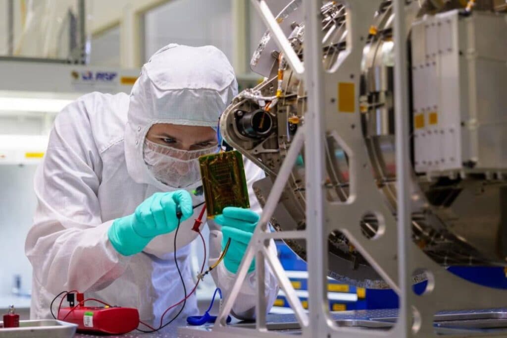 Person in a white jump suit and hood and mask leans over mechanical work