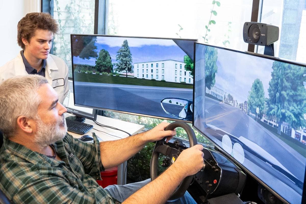 A man using a driving simulator while another man in a white lab watches him