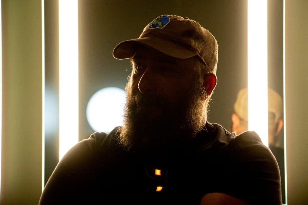 A headshot of a person with a hat and beard in dim lighting