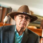 Rick Williams in a broad western hat, beaded hat band and feather quill, in a turquois shirt and grey suit jacket.