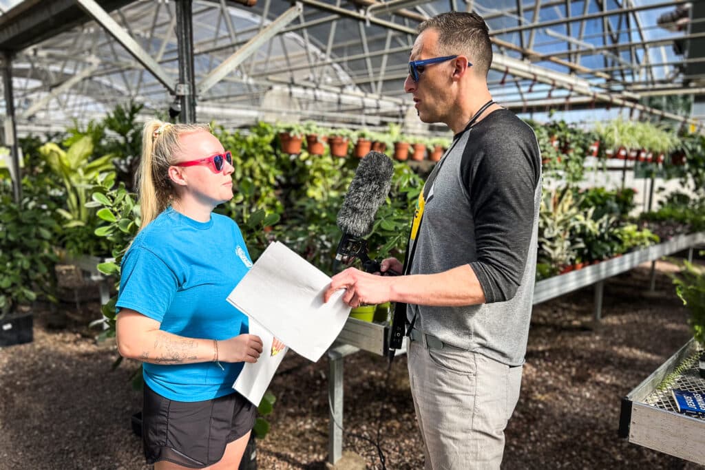 A woman in a blue shirt and sunglasses, and man in grey holding a microphone, talk for an interview inside a greenhouse at Phelan Gardens.