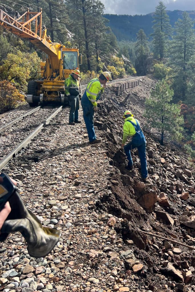 three men in yellow vests move rocks on a steep cliff near railroad.