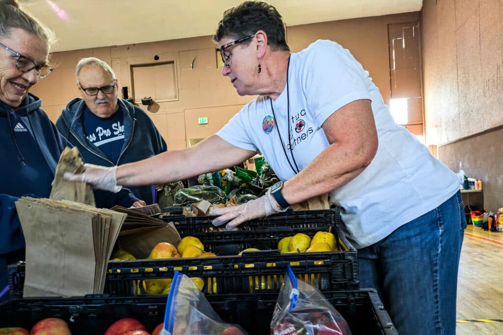 A woman in a white shirt denoting her as a volunteer with Mutual Aid Partners in Grand Junction hands a bag of produce to someone in line at a food distribution event inside a Grand Junction gymnasium.