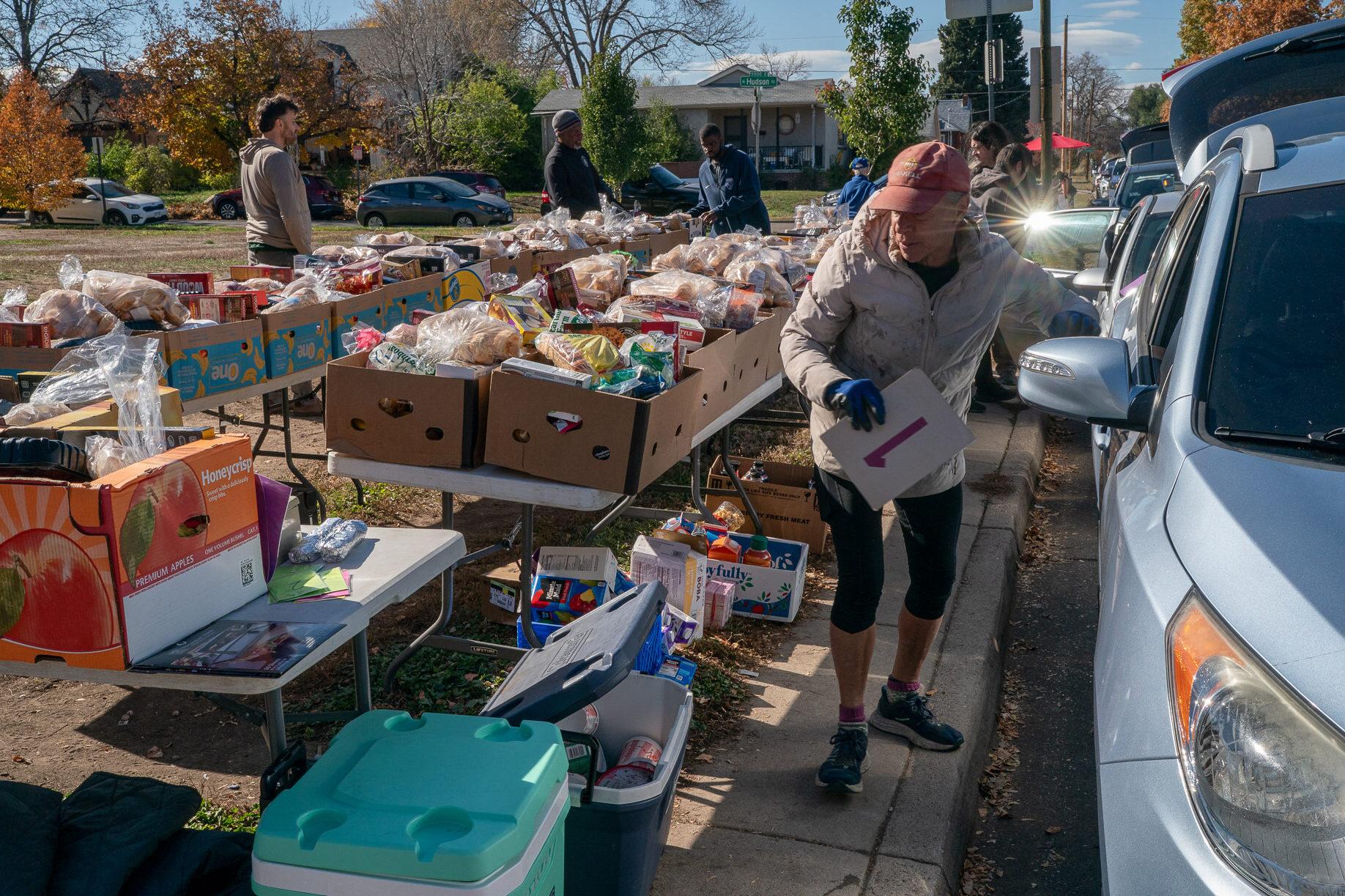 A person is standing next to a table filled with boxes of food, preparing to distribute the items. The table is located on the sidewalk, and there are several cars parked nearby.