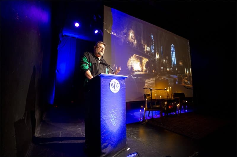 Photo shows a dark stage with a man at a podium in a black shirt with green alien heads and a black vest. To his right is a projection screen showing a fireplace in a living room in what looks like a stone manor.