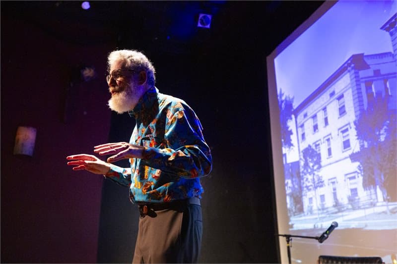 Photo shows a man in a blue long-sleeve shirt with a full beard and grey white air looking toward the left with his arms bent and hands extended. On the right is a large projection screen showing a historic building in Denver.