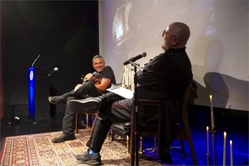 Photo shows two men in black seated on stage with their legs crossed. They have microphones in front of them and there's a white projection screen behind them.
