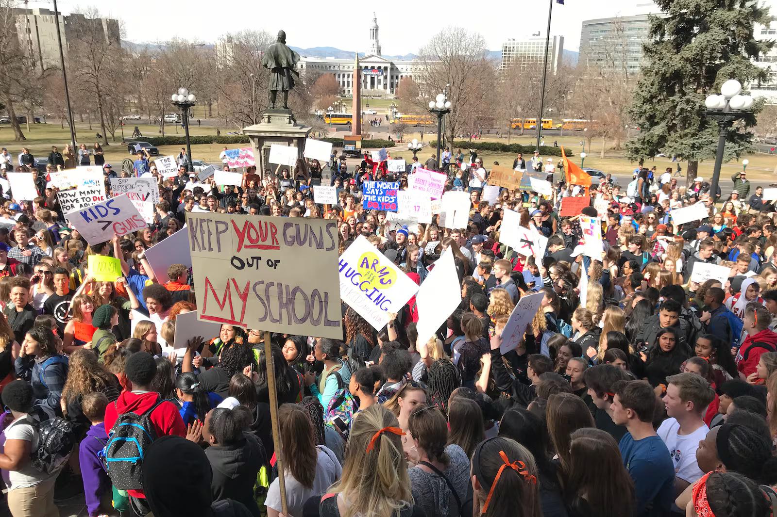 A large group of people gathered in a public space, holding signs and participating in a protest. They are advocating for gun control and school safety.