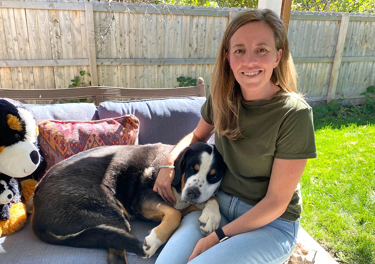A woman is sitting on a patio couch with a large brown and white dog resting its head on her lap. The woman is wearing a green shirt and is smiling at the camera.