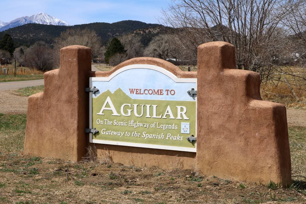A sign with pueblo-style architecture that says "Welcome to Aguilar" along 1st Street (County Road 63.1) in Aguilar, Colorado. The Spanish Peaks are visible in the background.