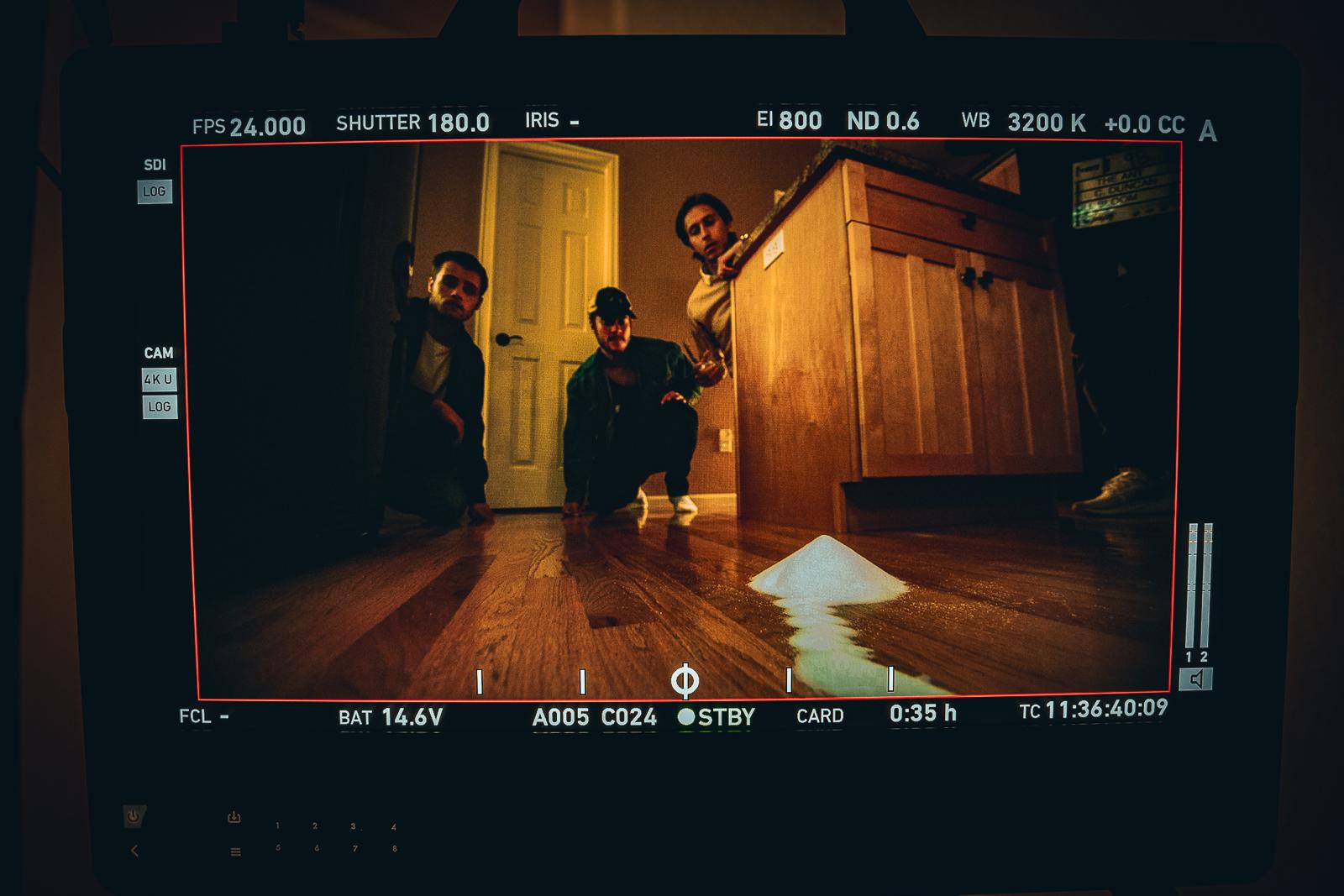 Image shows three people crouched on a wooden kitchen floor. One of the man is peering from behind a cabinet. The photo is framed in black with white camera markings.