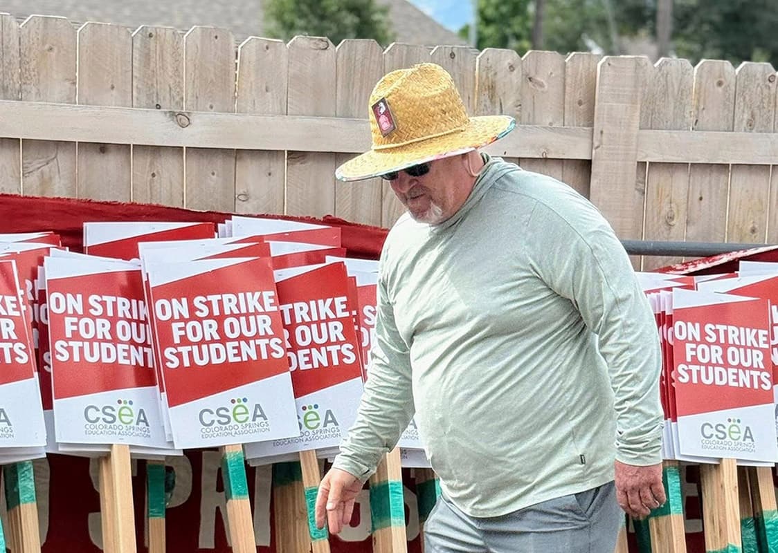 A man wearing a straw hat is walking past a row of signs. The signs read "On strike for our students" in bold capital letters.