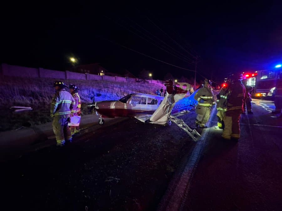 A small airplane with a bent wing sits on the side of a road, as five firefighters stand around it. The sky is dark, and emergency vehicle lights shine on the plane.