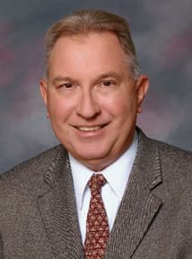 A man wearing a neutral colored suit, white shirt and tie is smiling for the camera.