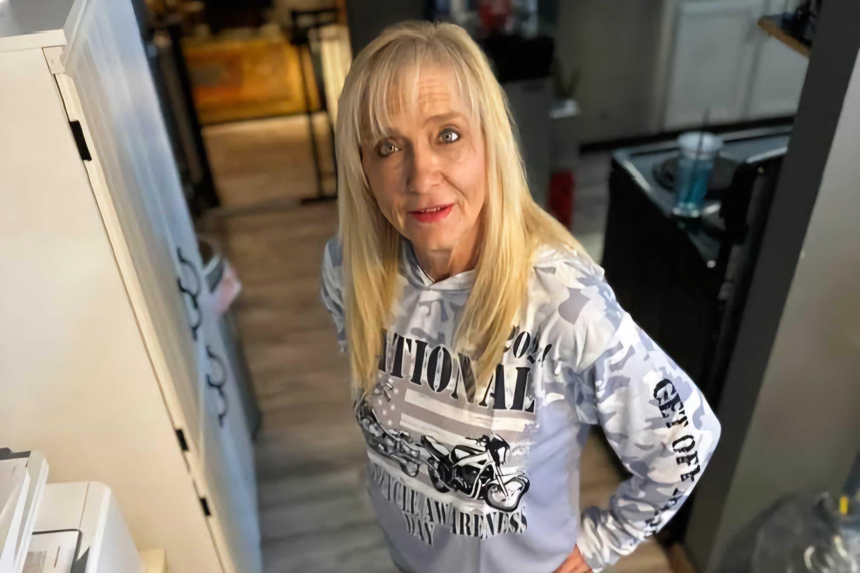 A woman with long blond hair is seen standing in a kitchen. She is wearing a grey and white sweatshirt with black lettering.