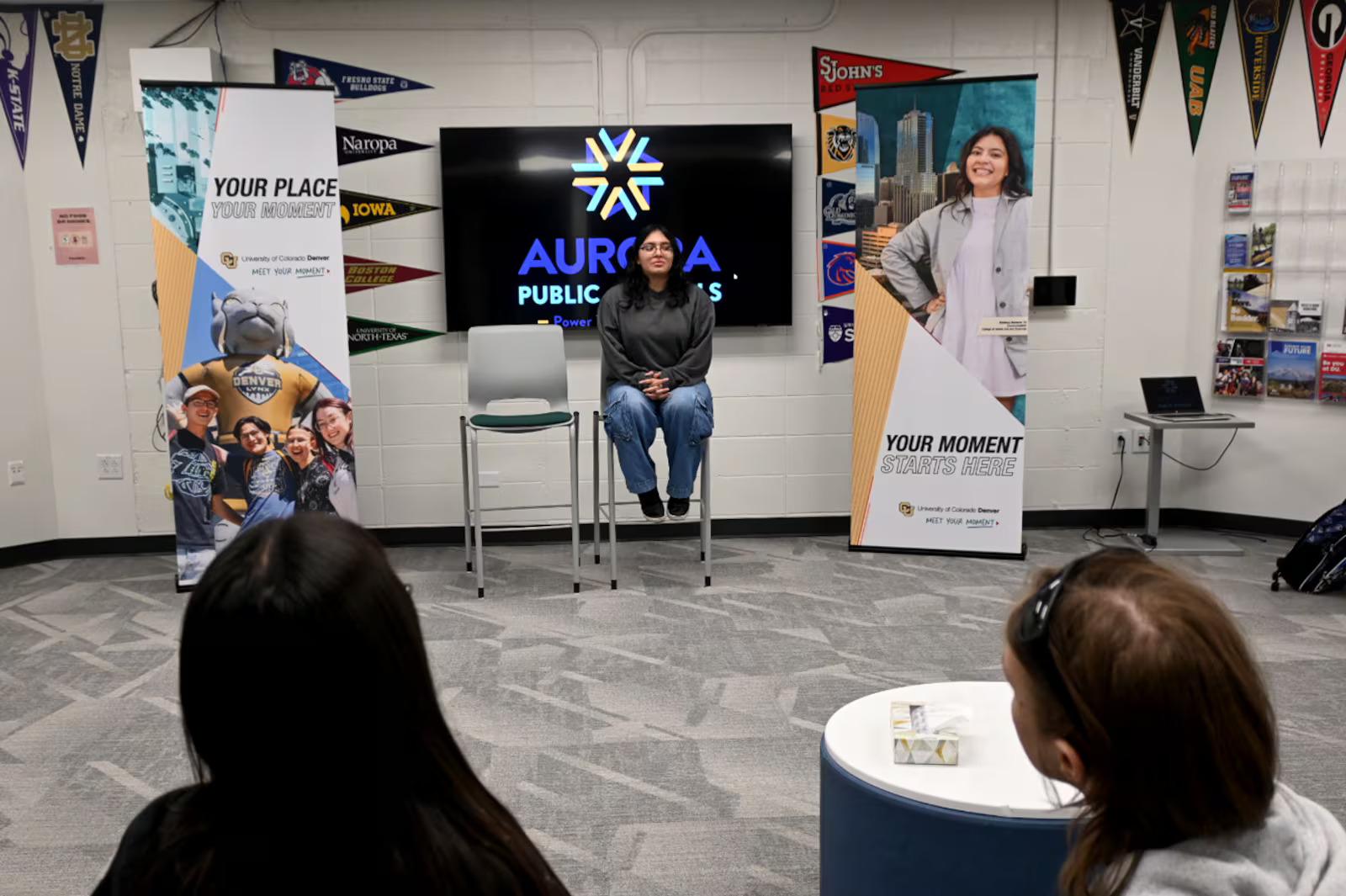 A person is sitting on a chair in front of a large screen, possibly discussing or presenting information. There are several other people in the room, some of whom are also sitting on chairs. The room appears to be a classroom.