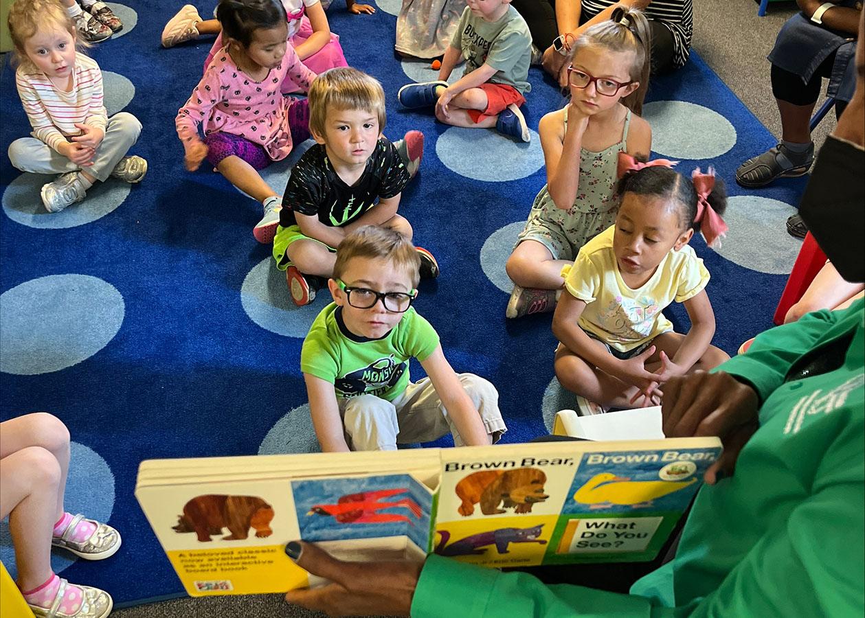A group of children is sitting on the floor, listening to a woman who is reading a book to them. The scene is set in a room with a carpeted floor, providing a comfortable environment for the children to sit and listen.