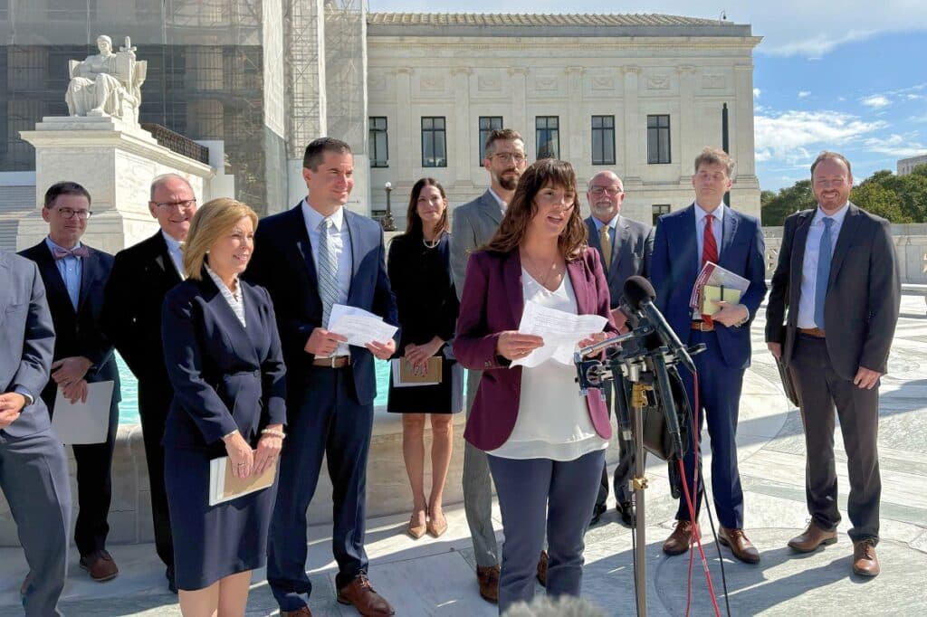 A group of people are standing in front of a building. They are standing behind a woman in front of a microphone.
