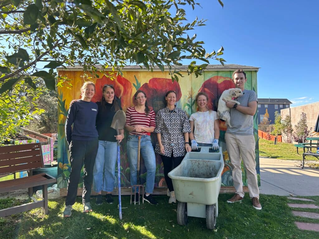 A group of people stand in front of the shed. One holds a shovel, another a pitchfork and one more a wheelbarrow.