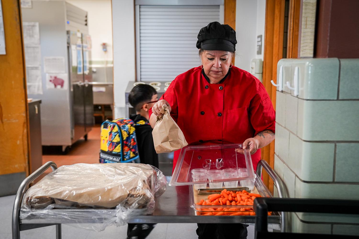 A woman dressed in a red professional-style kitchen shirt with a black cap holds up a brown lunch bag. Her left hand is lifting the lid off of a tray of baby carrots and young students with backpacks pass behind her.