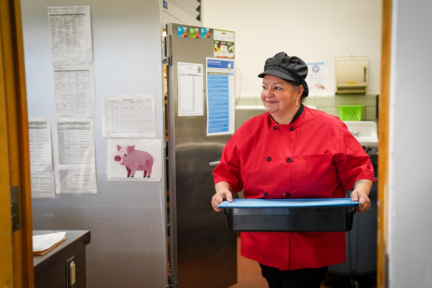 A woman dressed in a red professional kitchen-style button down shirt and a black hat carries a tray of food from the fridge to a counter. She smiles and looks off to her right.