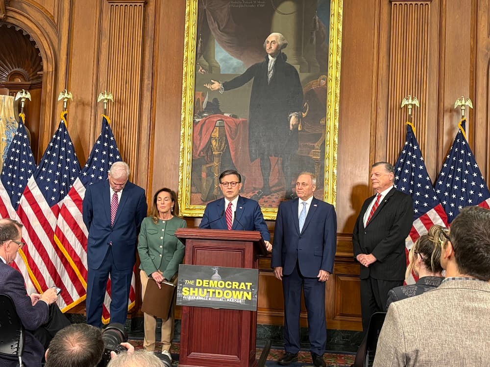 Speaker Mike Johnson stands at a podium with a sign that reads "the Democrat shutdown." He is flanked by other Republican Representatives, with a backdrop of flags and a portrait of George Washington.