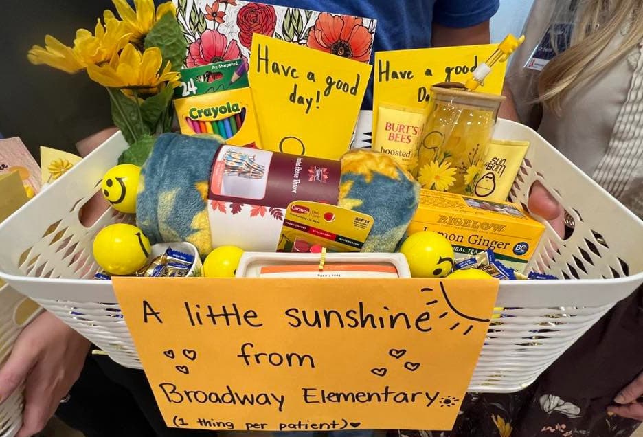 A basket filled with various items, including a card that says "A Little Sunshine from Broadway Elementary." The basket is placed on a table, and there are multiple books and a vase in the scene.