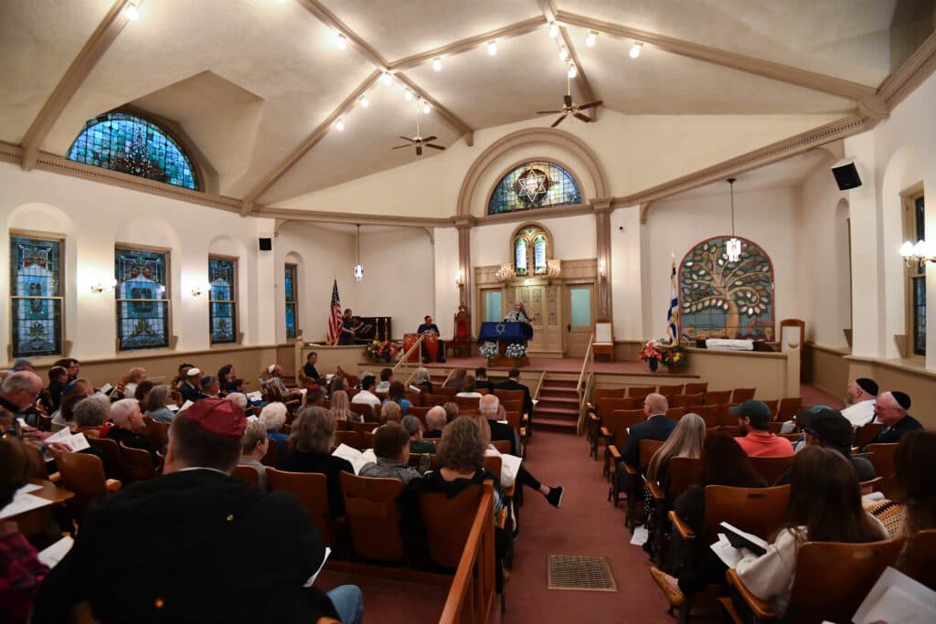 Most of the seats are filled in a synagogue sanctuary with stained glass windows and other Jewish motifs