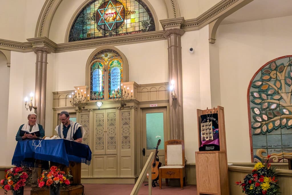 Two men wearing prayer shawls and skull caps stand beneath colorful leaded glass windows at a lectern covered with blue velvet