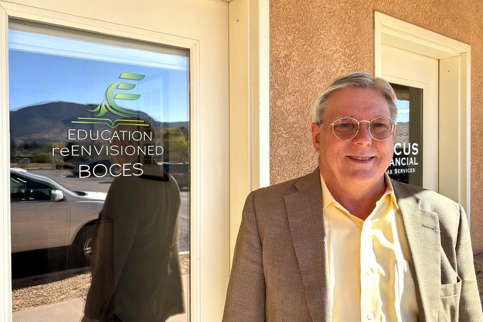 A man in a tan jacket is standing in front of a door with a window. The door has a sign on it that says "Education Invisioned."