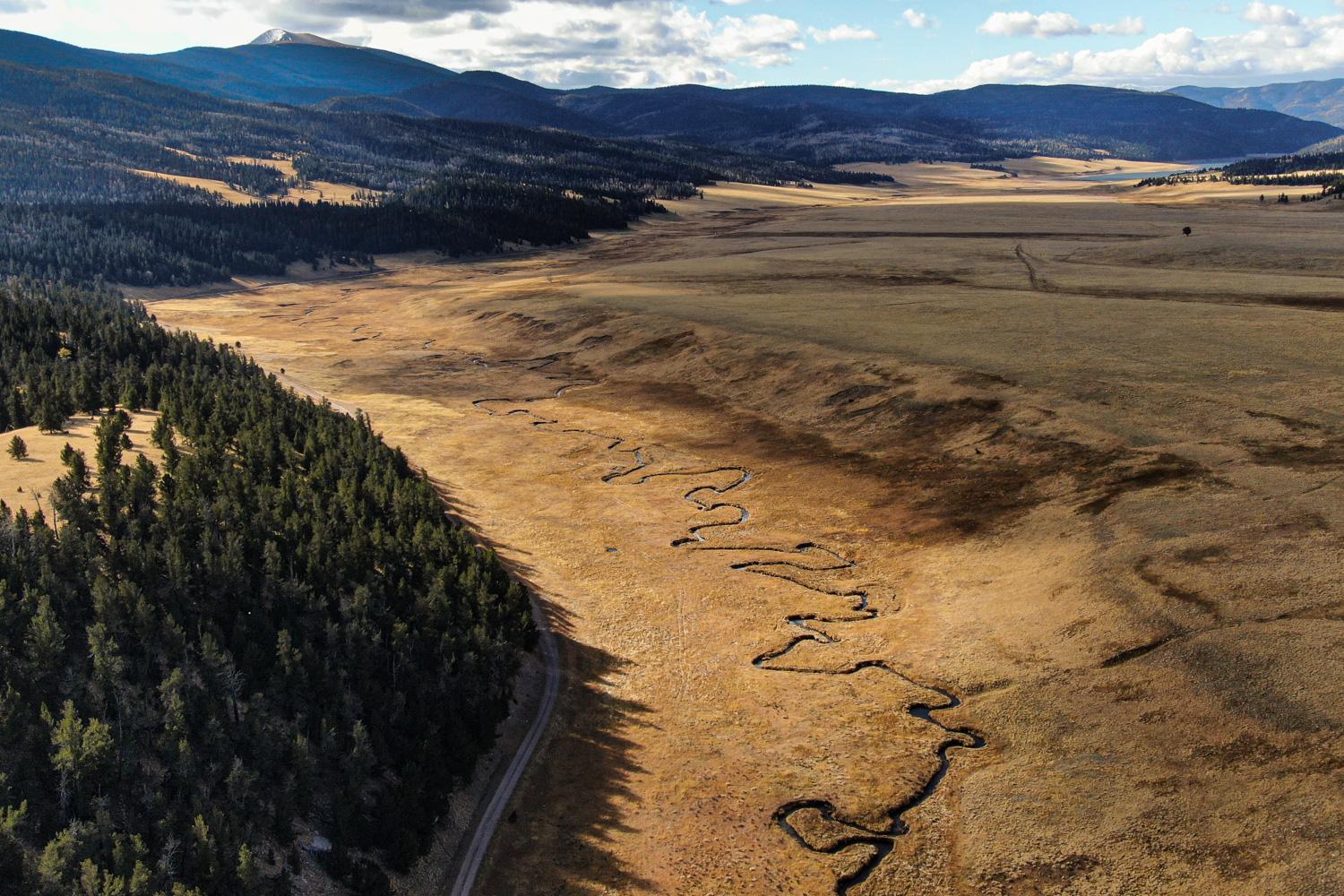 Large grassland area with a creek and a road running through it. The field is surrounded by tree covered mountains. The entire scene appears to be empty, with no visible vehicles or people.