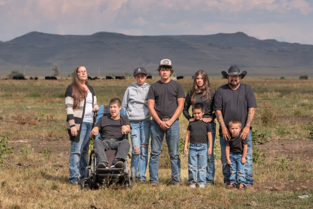 A group of people, including both adults and children, are posing for a picture in a field. They are standing in front of a hill, and there are cows in the background.