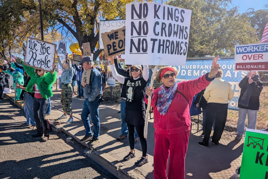 A group of people are gathered on a street, holding signs and protesting. They are standing in front of a fence. The signs they are holding are visible, and they appear to be advocating for a cause.