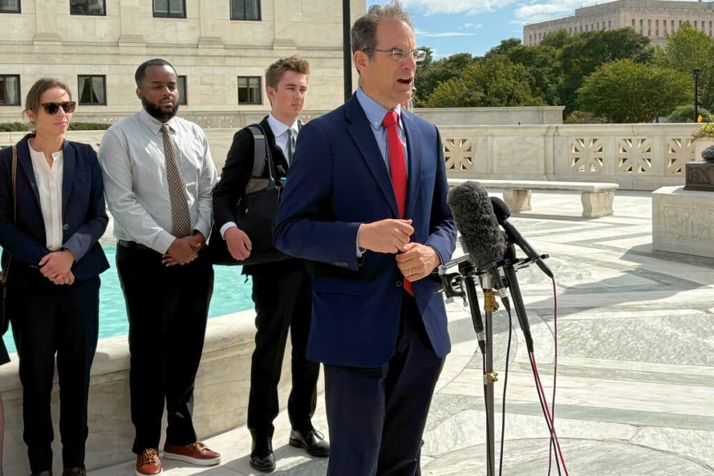 A man in a suit is speaking into a microphone, with three other people standing behind him.