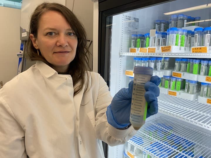 A woman in a lab coat is holding a vial of green liquid. She is standing in front of a refrigerator, which is filled with various bottles. The woman appears to be a scientist or researcher, possibly working on a project involving the green liquid.