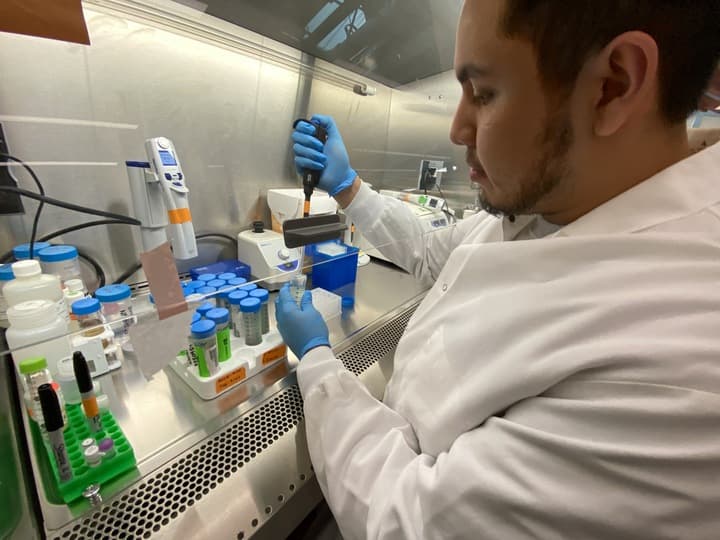 A man in a lab coat is working on a table with various items, including a microscope, a cell phone, and a computer. He is wearing gloves and appears to be handling a sample. The scene suggests that the man is conducting scientific research or experimentation.