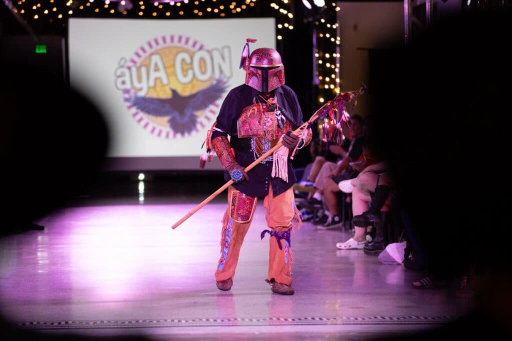 A model in a an orange and black outfit wearing a helmet and holding a long pole