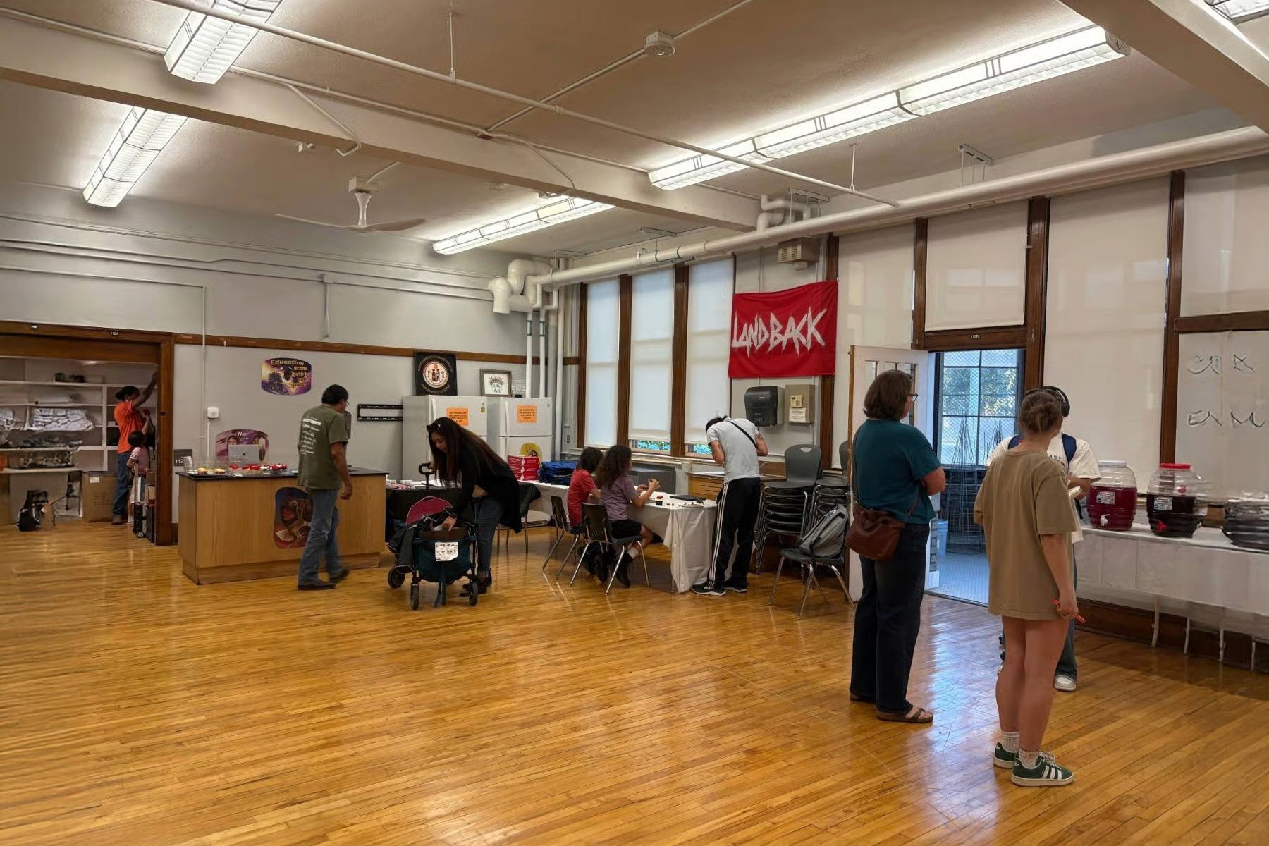 The image depicts a group of people gathered in a community center. There are several chairs and tables scattered throughout the space.