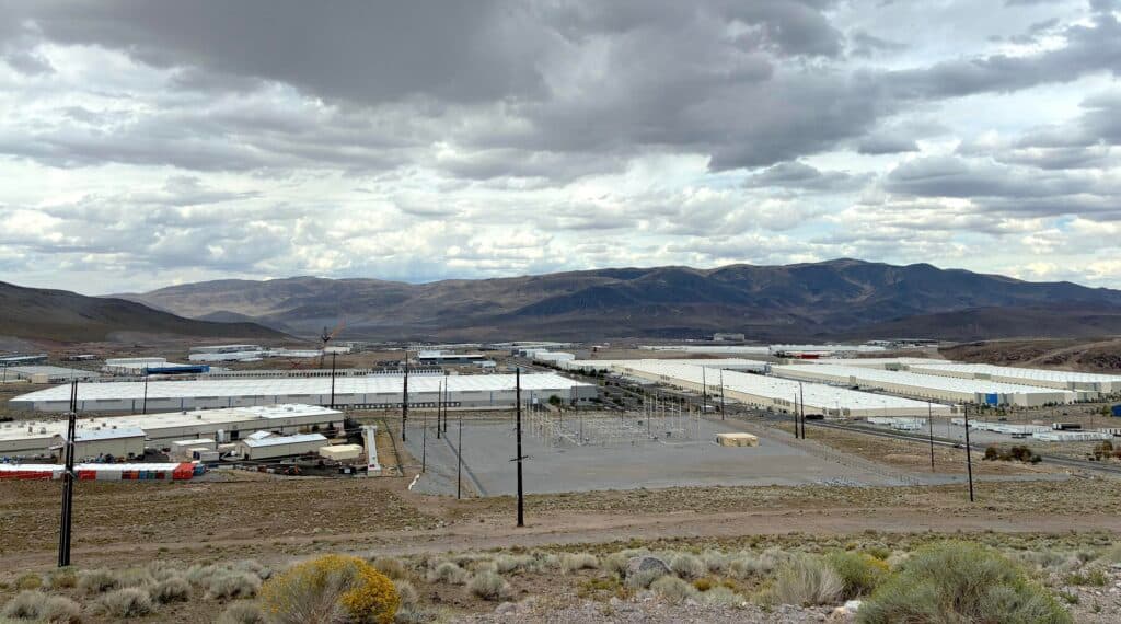 This is a landscape image overlooking a large industrial park filled with long, flat-roofed buildings used for manufacturing, warehousing and data centers. The sky is filled with clouds hovering over rolling green-brown mountains.