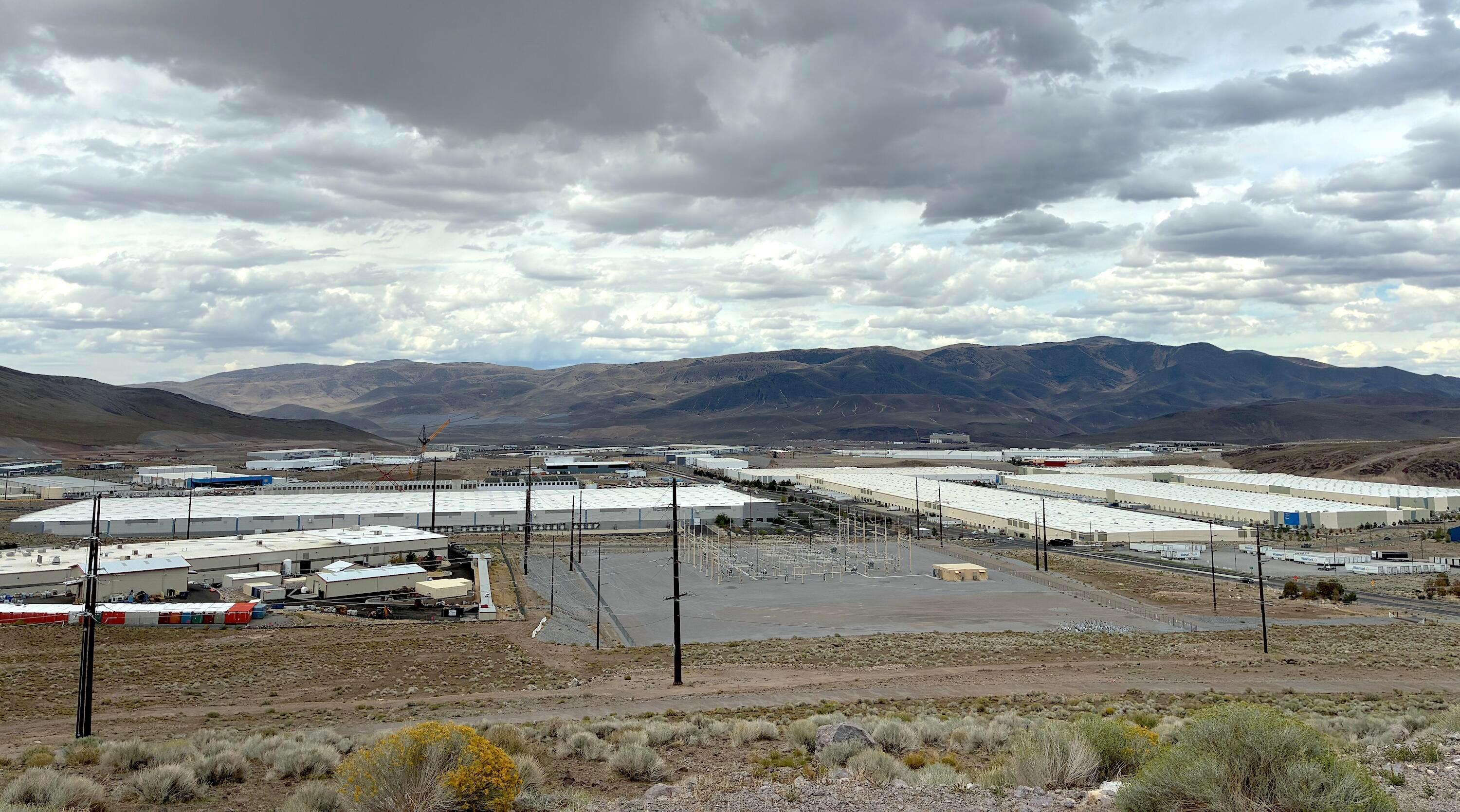This is a landscape image overlooking a large industrial park filled with long, flat-roofed buildings used for manufacturing, warehousing and data centers. The sky is filled with clouds hovering over rolling green-brown mountains.