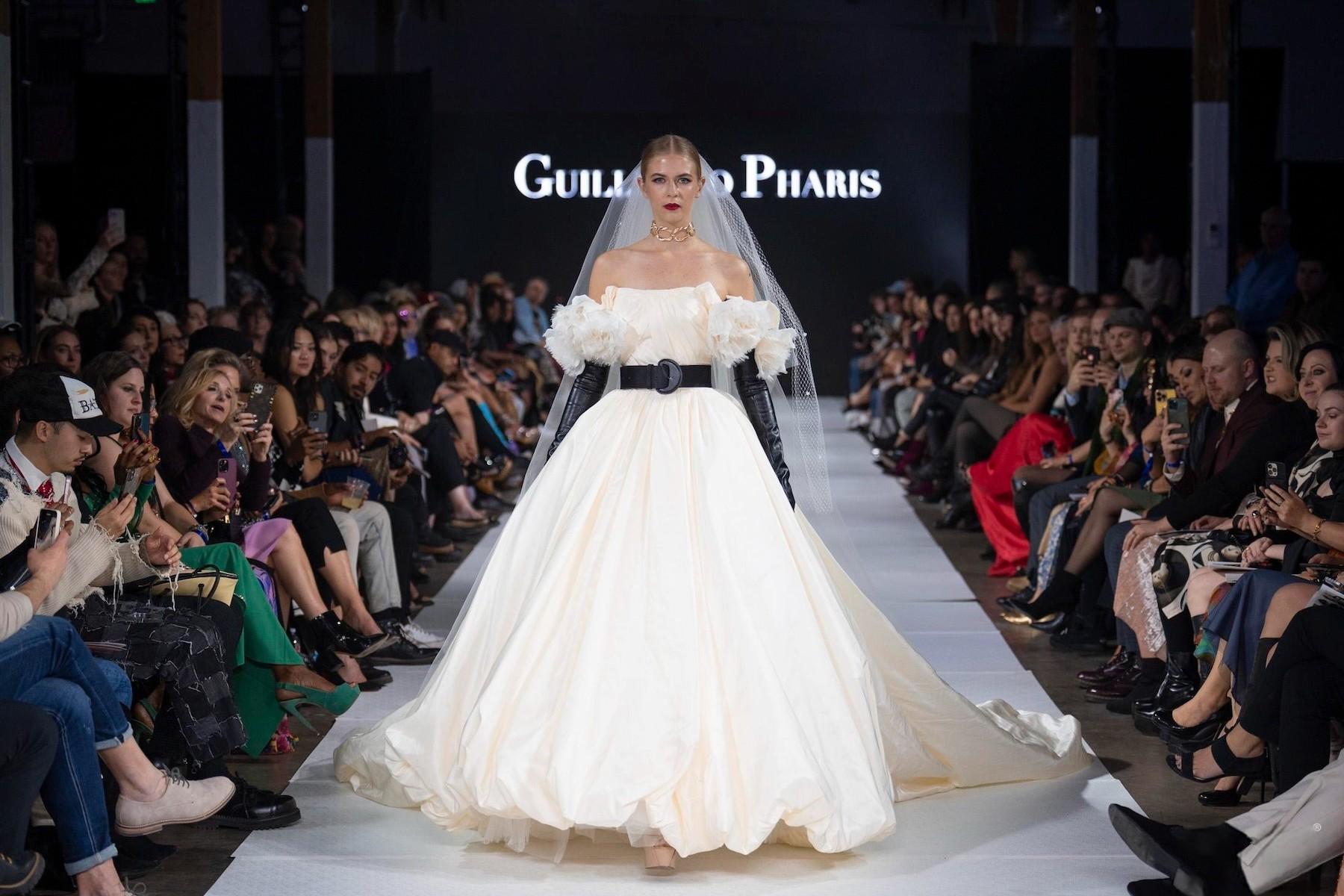 A model in a large, off-white gown with a black belt and tall black gloves walks the runway.