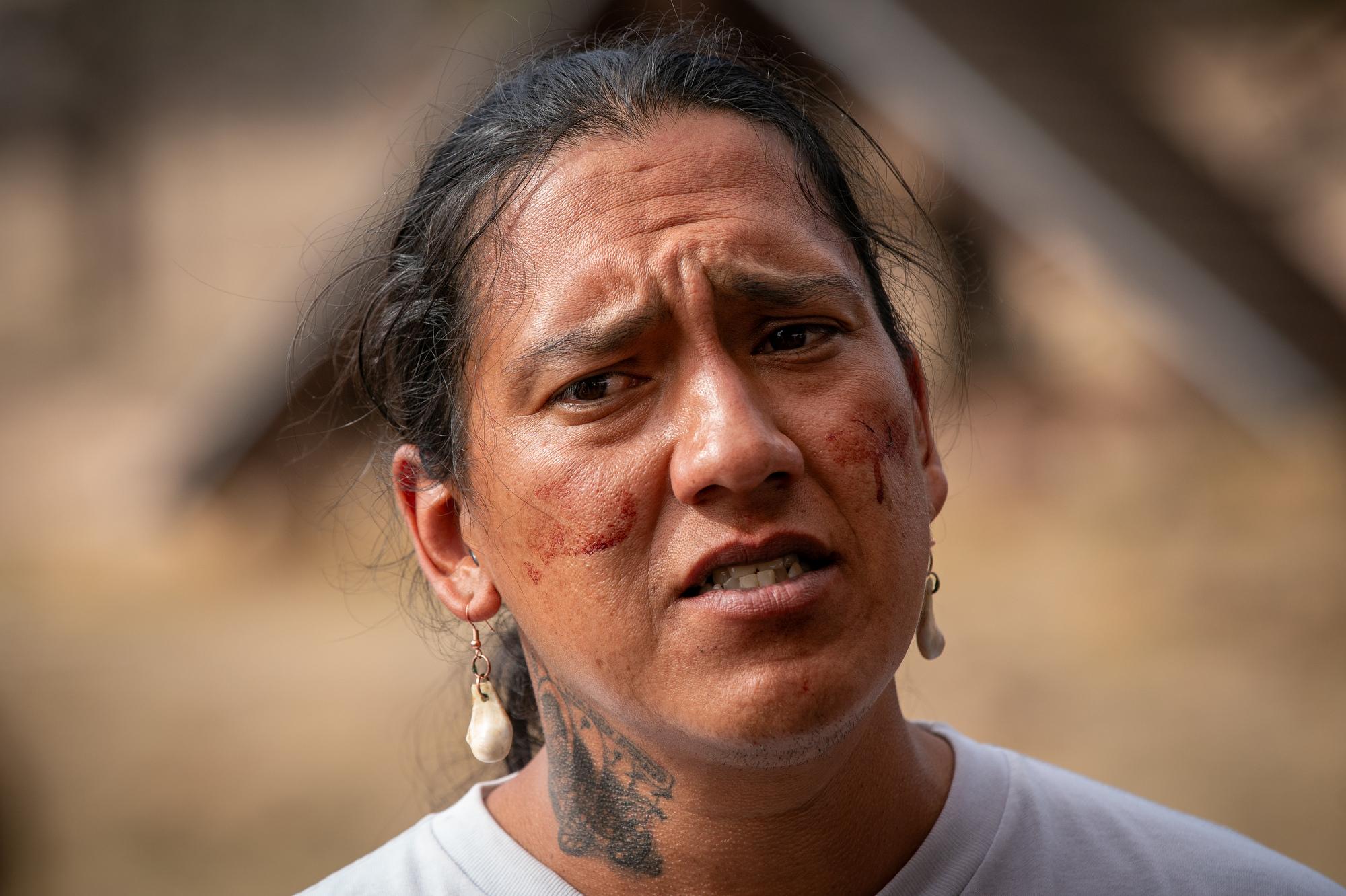 A man with earrings and a tattooed neck is looking down and to the right of the camera. He is wearing a white shirt.